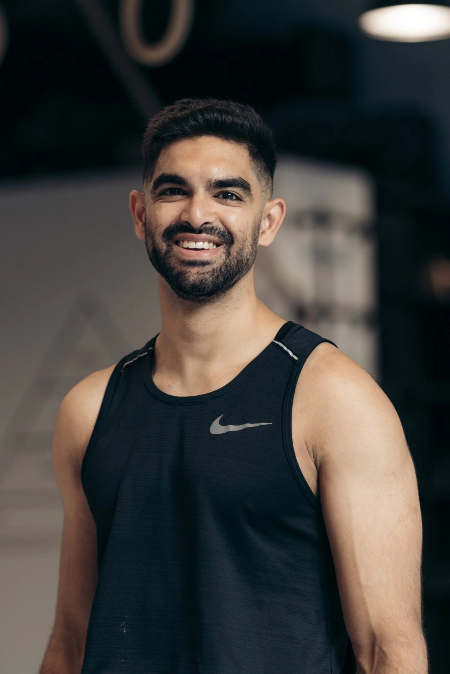 A man with dark hair and a beard smiling, wearing a black Nike sleeveless athletic shirt, in a gym or fitness center.