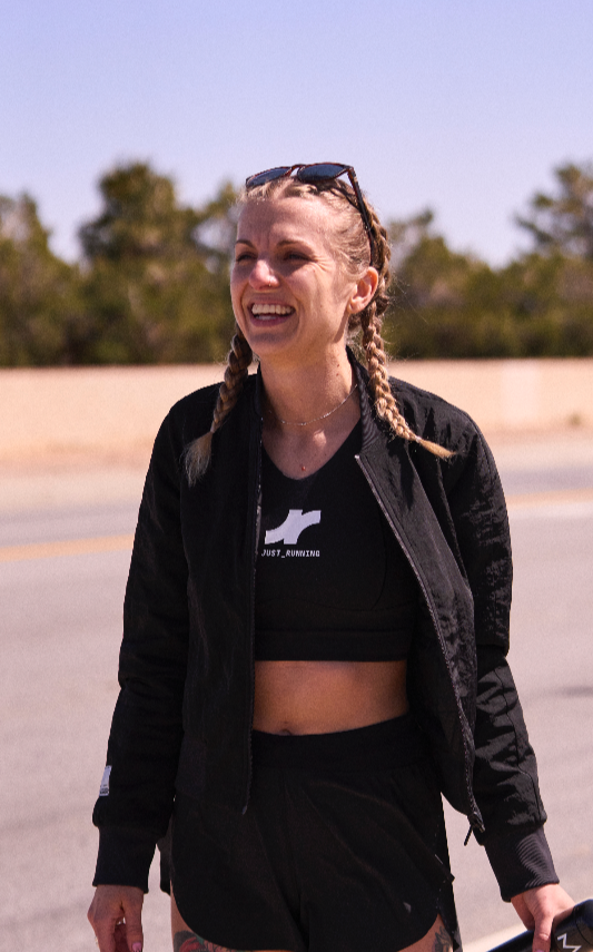 Young woman with braided hair smiling outdoors on a sunny day, wearing a black cropped top, black jacket, and sunglasses on her head.