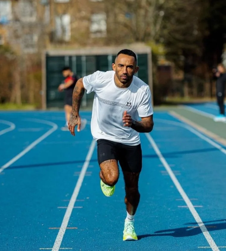 A man running on a blue outdoor track during daytime, wearing a white shirt, black shorts, and yellow running shoes, with trees and a fence in the background.