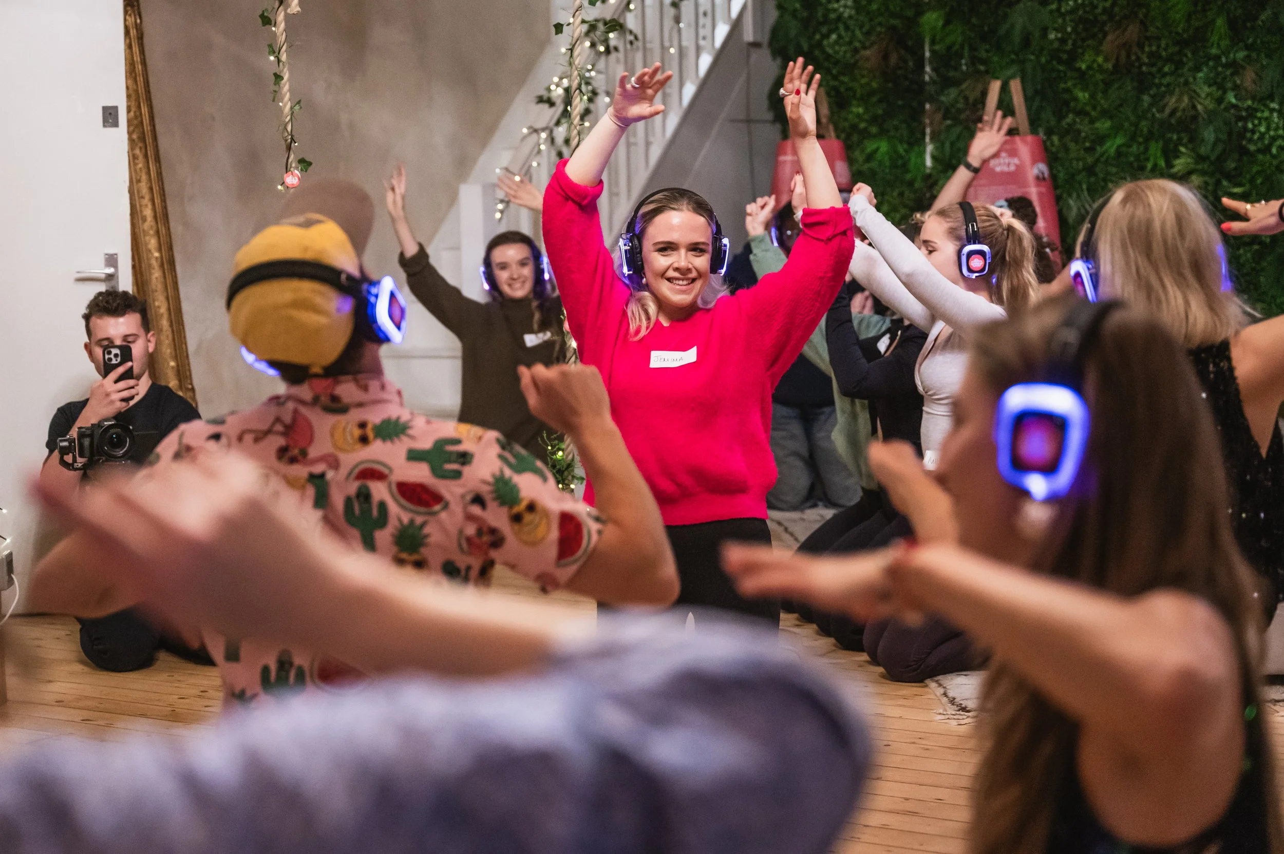 People dancing and celebrating at a festive party, some wearing headphones and colorful clothing, with a decorated wall and greenery in the background.