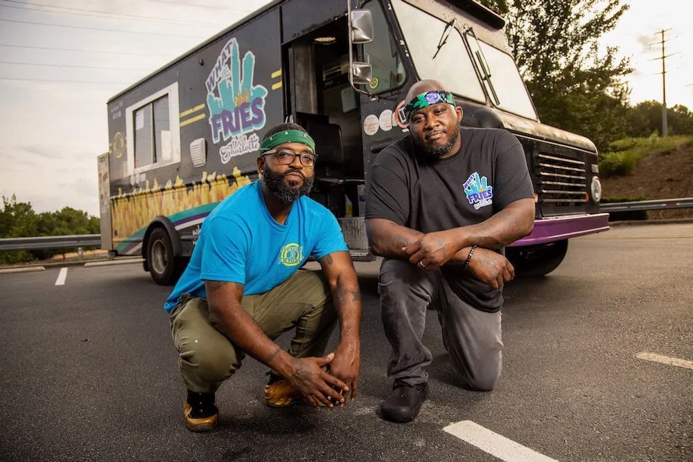  Photo:  Two men (food truck owners) kneeling in front of their food truck, while wearing matching headbands and logo shirts.  