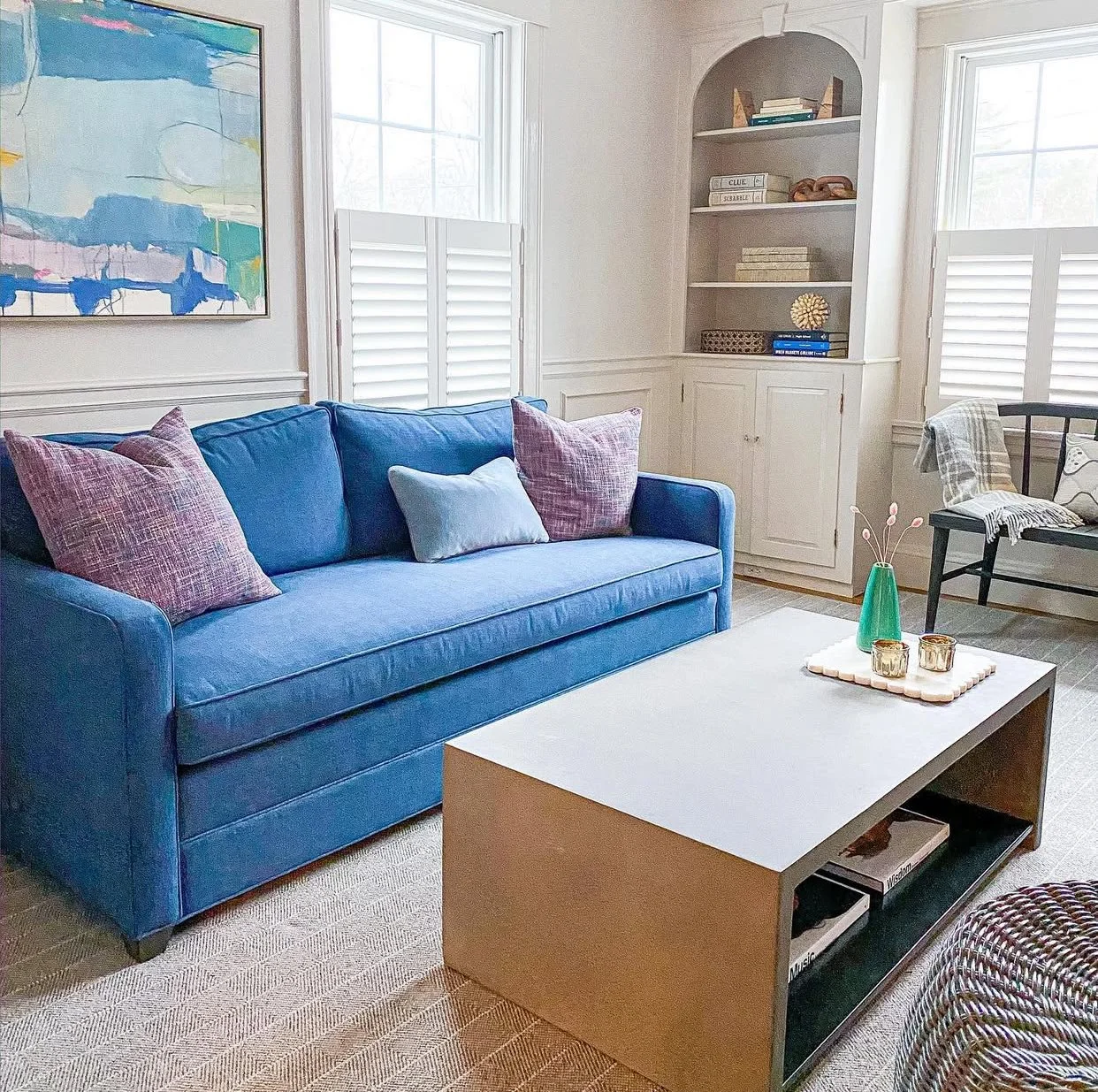 Living room with a blue sofa, purple and light blue pillows, a white coffee table with decorative items, a black chair with a throw blanket, a built-in white bookshelf, and windows with white shutters.