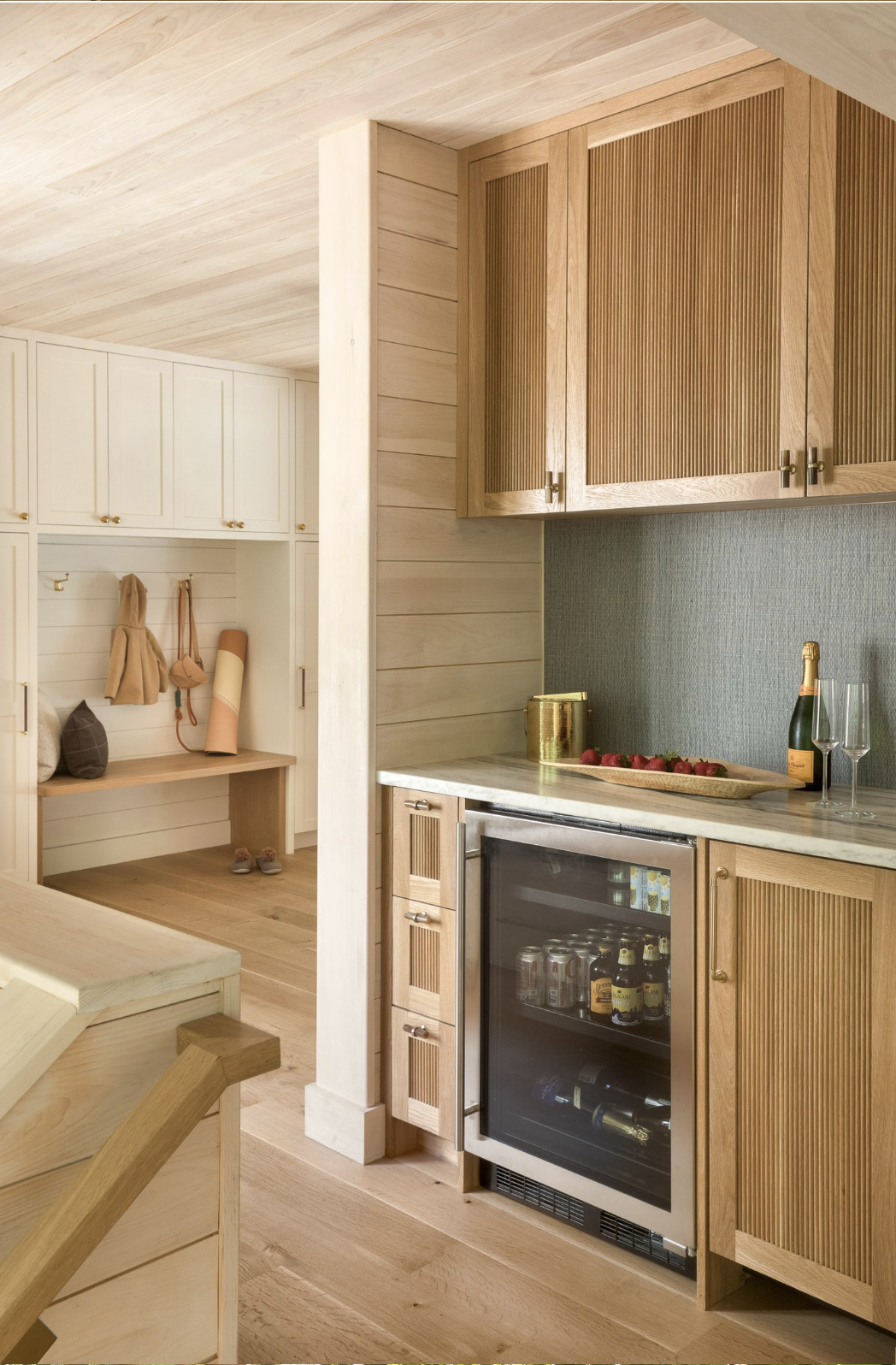 A cozy kitchen and entryway with wooden cabinetry and furniture, a countertop with a tray of strawberries, champagne bottle, and glasses, and a small drink refrigerator below the counter.