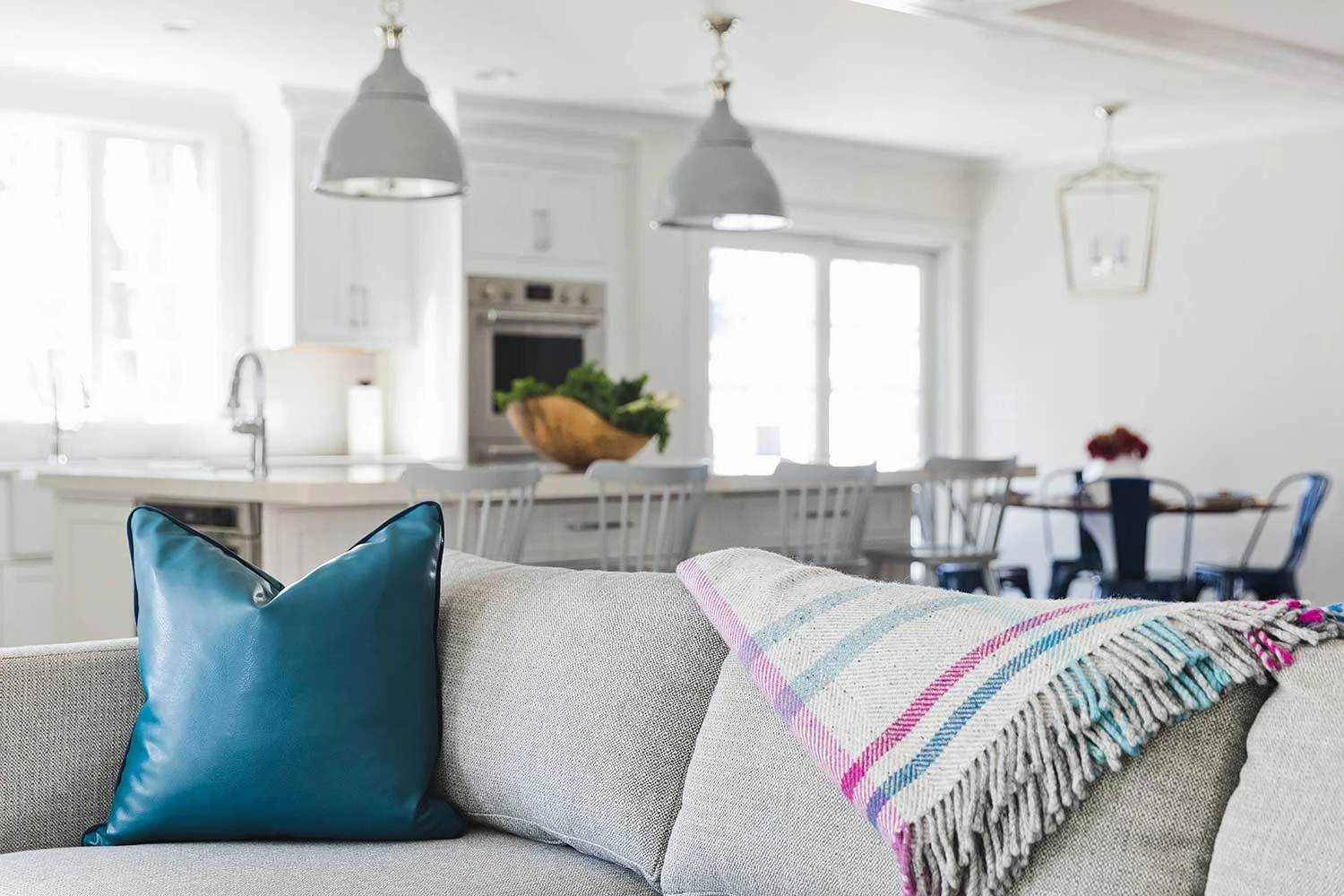 Living room with beige sofa, blue leather pillow, and pink striped throw blanket, overlooking a white kitchen and dining area with pendant lights and large windows.