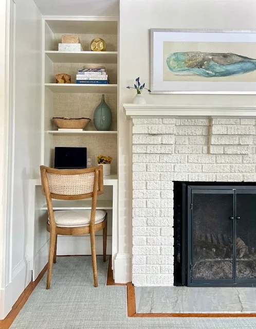 A small home office nook with a built-in white bookshelf next to a white brick fireplace with a black screen. The bookshelf holds decorative objects, books, a green vase, and a small laptop or tablet. A wooden chair with a woven back and white cushio