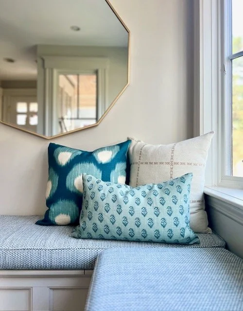 A cozy window nook with three decorative pillows, a mirror on the wall, and sunlight streaming through a nearby window.