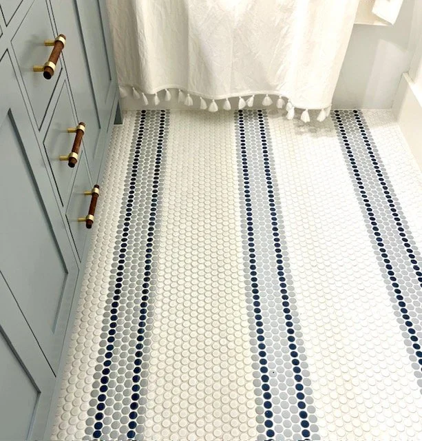 Bathroom floor with white and dark blue penny tiles, blue dresser with gold handles on the left, and white curtain with tassels at the top.