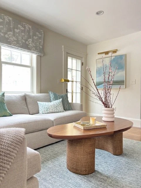 Living room with a beige sectional sofa, decorative pillows, a wooden coffee table with a vase of twigs, a wall painting, and windows with floral curtains