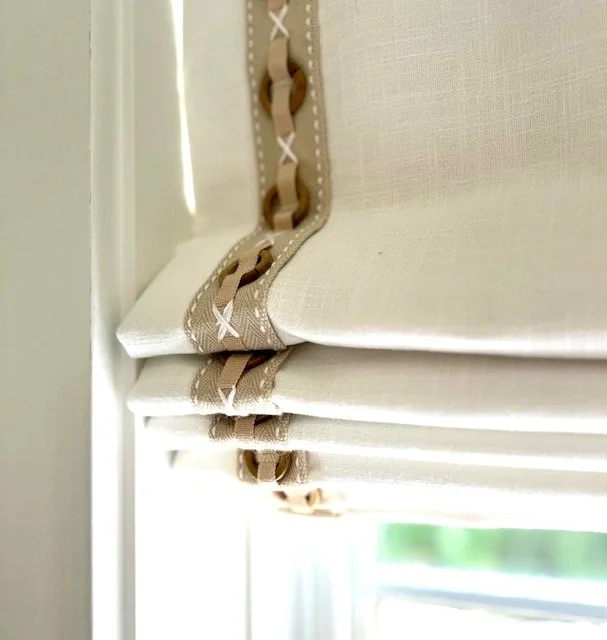 Close-up of beige Roman window shade with decorative trim, partially lowered in front of a window.