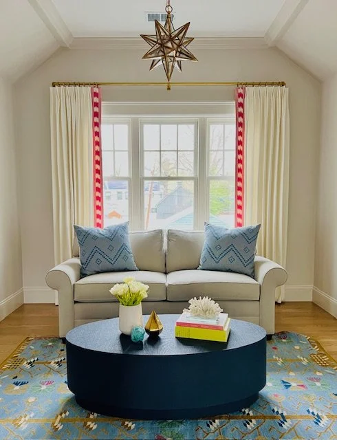 Living room with a white sofa, blue pillows, a black oval coffee table with decorative items, a colorful rug, and cream curtains with red and white patterned trim. A star-shaped pendant light hangs from the ceiling in front of a window with a view of
