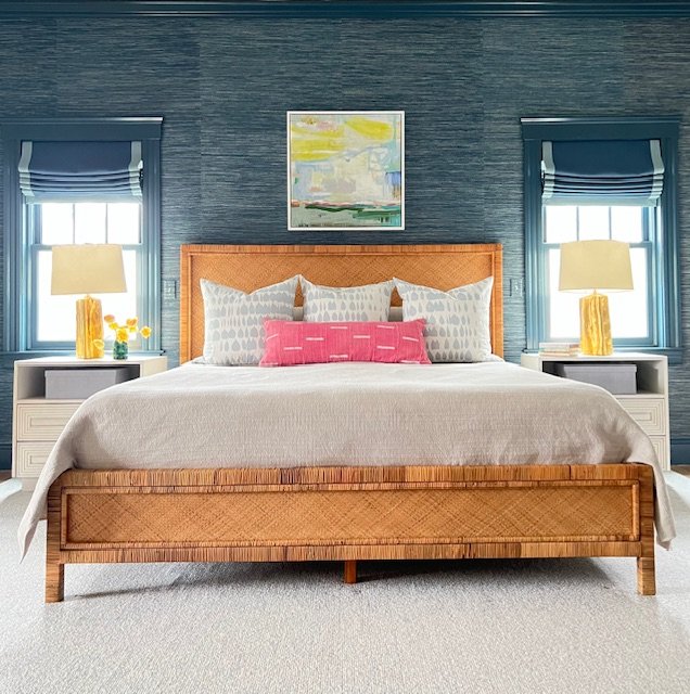 A bedroom with a wooden bed frame, beige bedding, decorative pillows, two nightstands with lamps, and a colorful abstract painting on a textured blue wall.