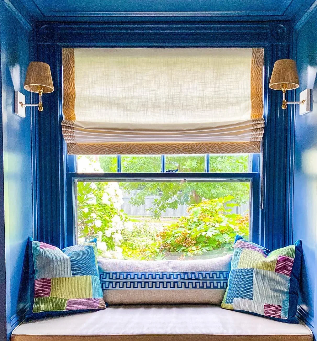 A window seat with blue walls, two patterned pillows, a cushioned bench, a beige Roman shade, and a view of greenery outside