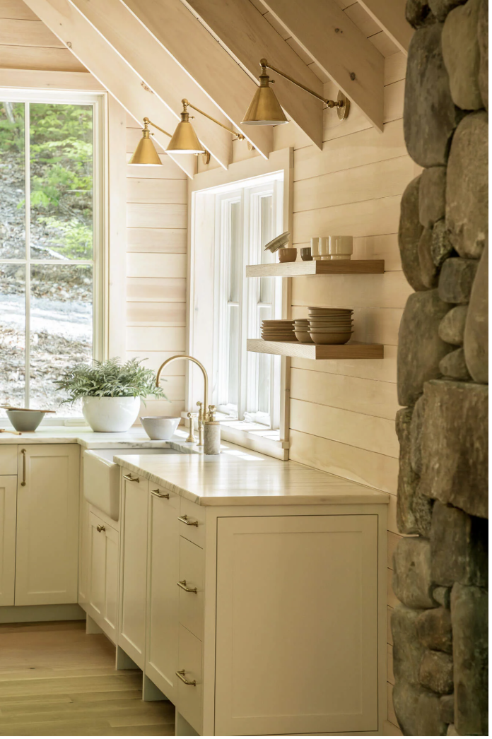 Kitchen with cream-colored cabinets, a window with a scenic view, open wooden shelves with dishes, brass lighting fixtures, a potted plant, and a stone wall on the right.