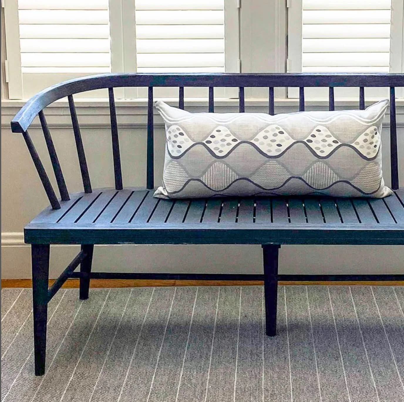 A dark wooden bench with slatted seat and backrest, placed in front of white window shutters, with a decorative rectangular pillow with wavy lines and polka dots resting on it.