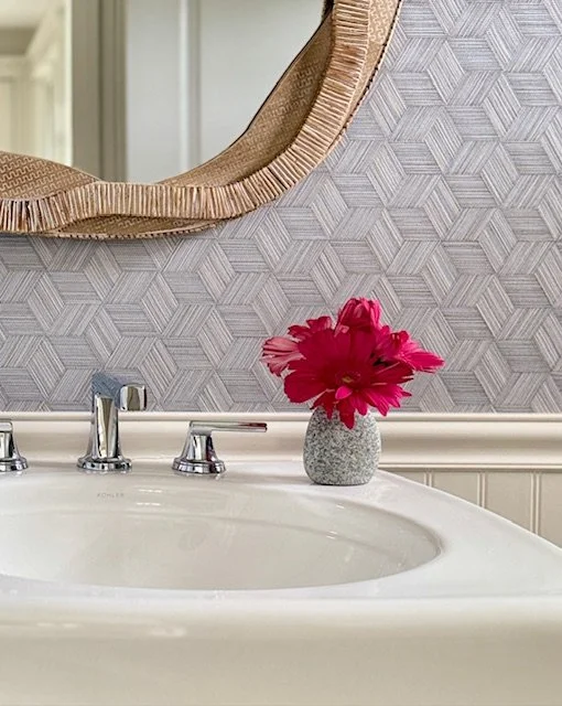 Bathroom sink with a vase of bright pink flowers, mirror, and textured wallpaper in the background.