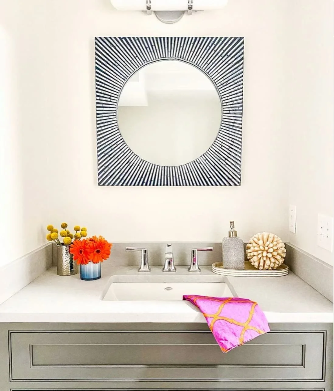 Bathroom vanity with a white countertop, gray cabinet, and colorful decor. There is a square mirror with black and white geometric rays hanging above. A pink and orange towel is draped over the sink. Decor items include yellow pom-pom flowers in a si