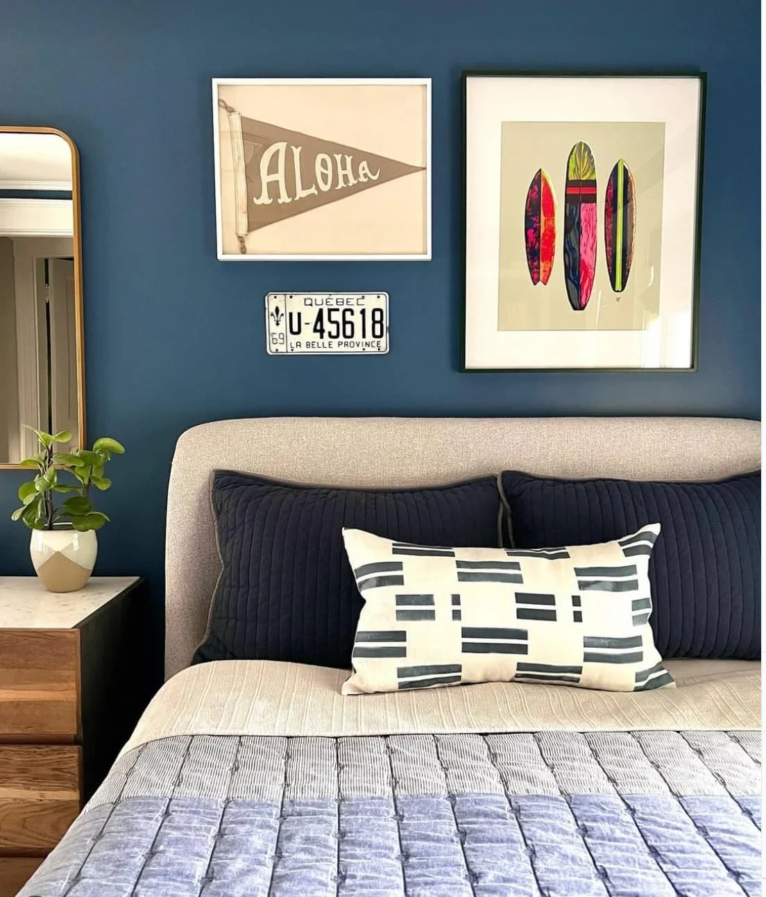 A bedroom with a bed that has a beige headboard, dark blue quilted pillows, and a black and white patterned bolster pillow. The wall behind the bed is painted blue and decorated with framed art, a vintage Quebec license plate, and a small mirror. The