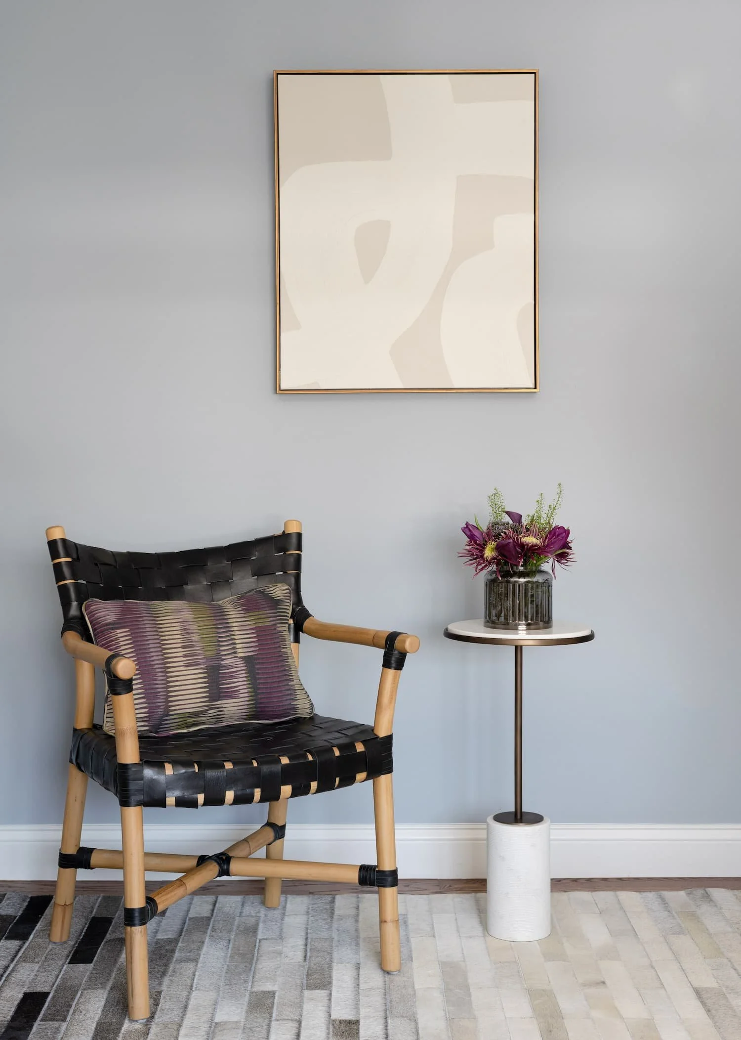 A modern interior corner with a black woven leather and wood armchair, a small round side table with a floral arrangement, and an abstract artwork on a light gray wall.