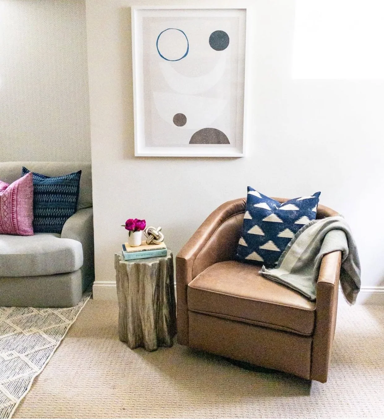 A cozy living room corner with a beige armchair, a blue cushion with a white triangular pattern, a throw blanket, a wooden side table with pink flowers in a white vase, and a framed abstract art piece on the wall.