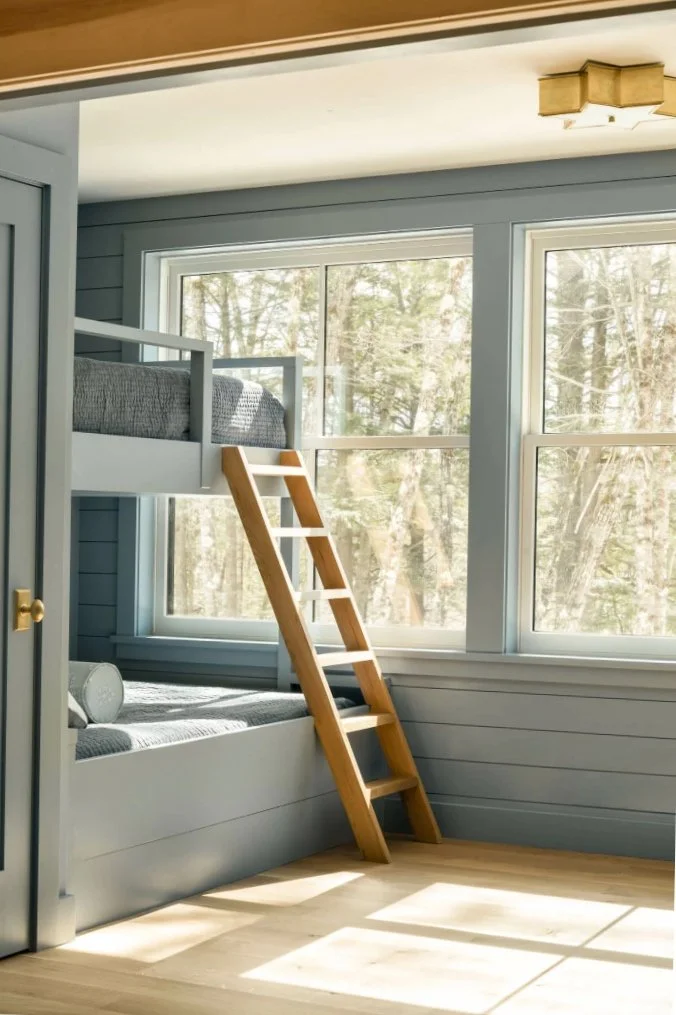 A cozy bedroom with a built-in bed and a loft bed accessed by a wooden ladder, large windows with a view of trees outside, light wood flooring, and soft natural lighting.