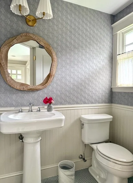 Bathroom with a wall-mounted mirror, a white pedestal sink with a small pink flower in a vase, a white toilet, a small trash can, a window with white trim, and patterned gray wallpaper.