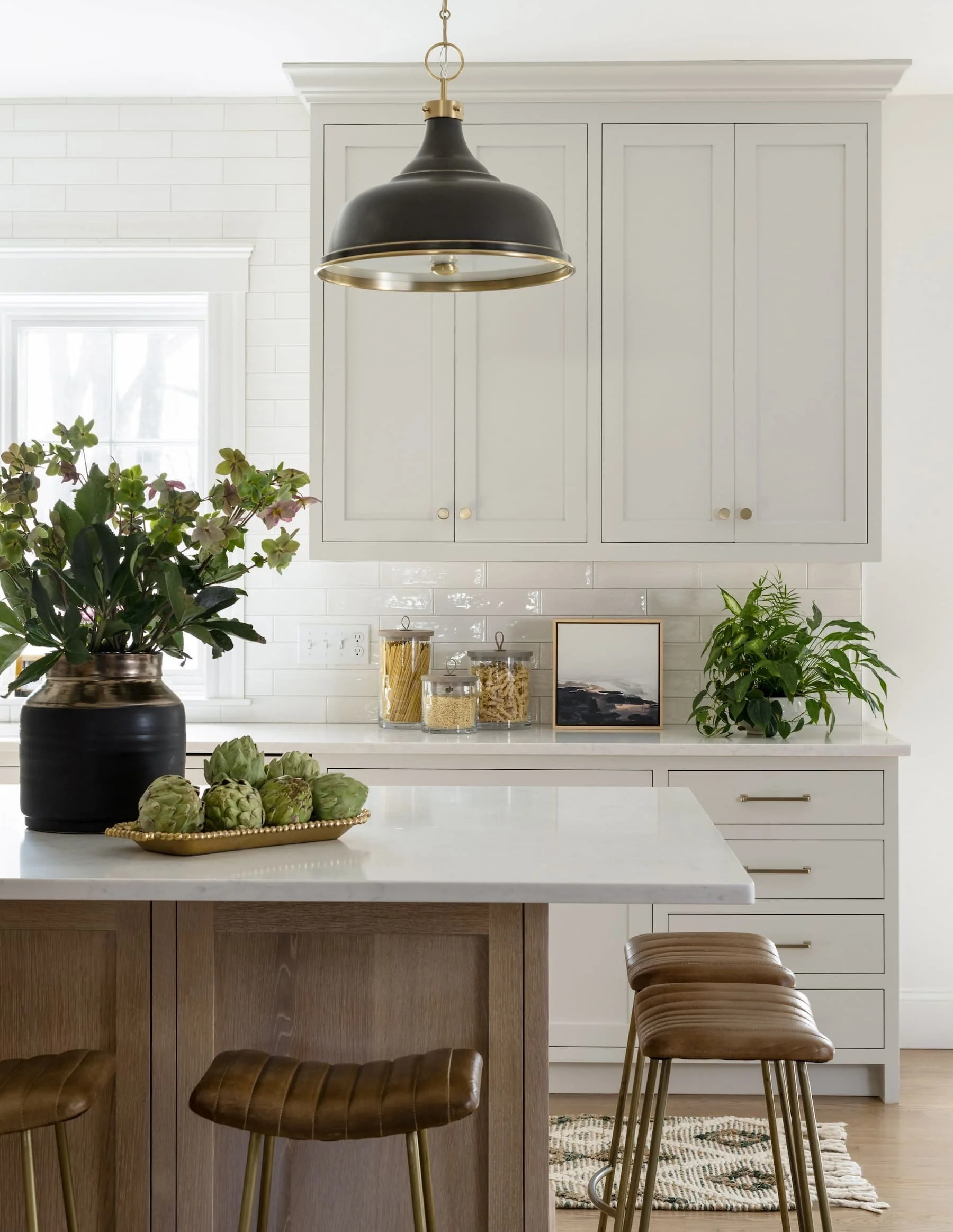Modern kitchen with white cabinets, a white countertop, and a kitchen island with wooden stools. Decor includes plants, art, and jars of pasta and grains, with a black hanging light fixture.