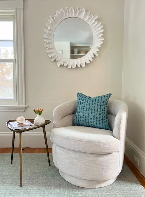 A cozy corner in a room with a round white armchair, blue patterned throw pillow, small side table with a vase of flowers and a book, a wall mirror with a decorative frame, and a window with natural light.