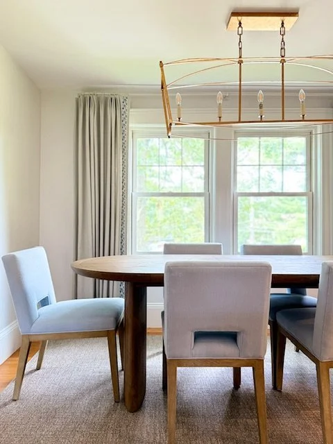 Dining room with a round wooden table, upholstered chairs, and a chandelier hanging from the ceiling. Large windows with white curtains let in natural light.