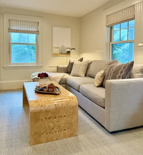 Living room with a beige sectional sofa, several pillows, two windows with blinds, a beige textured coffee table with decor, a framed picture on the wall, and a white wall with a small lamp.