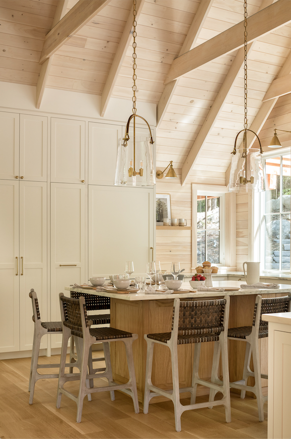 A cozy kitchen with a high, sloped wooden ceiling, white cabinets, a center island with a marble top, and six seating chairs. Large windows allow natural light in, and pendant lights hang from the ceiling.