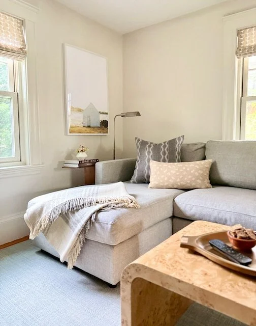 A cozy living room with a light gray sectional sofa, two decorative pillows, a small wooden side table with a floral arrangement, a wall-mounted lamp, a framed landscape picture, and two windows with beige Roman shades.