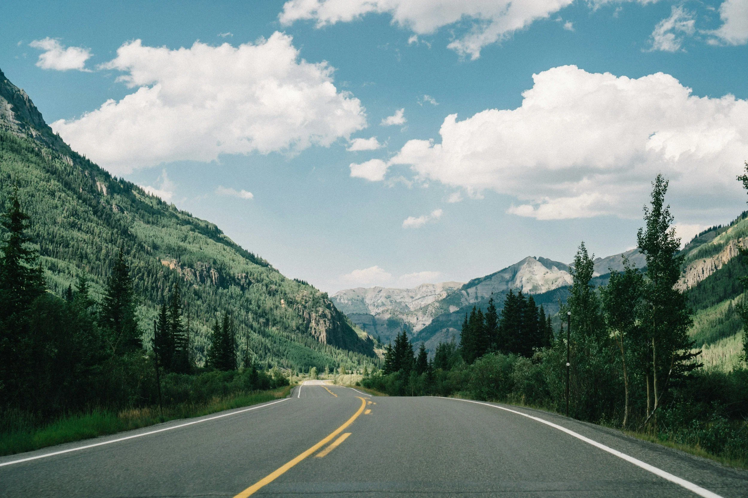 A scenic mountain road with lush green trees on either side, winding through a mountainous landscape under a partly cloudy sky.