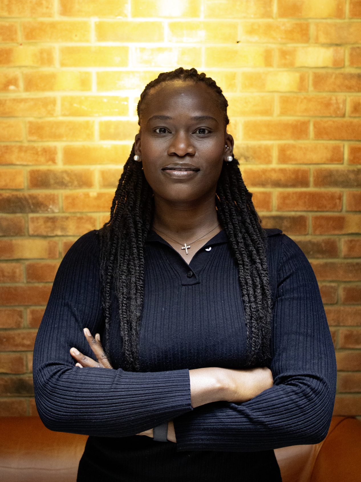 A female founder looks down the lens of the camera with her arms folded, the lighting providing a warm halo from behind her head, against the brick wall behind