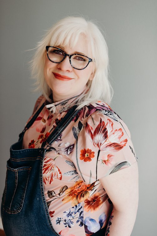 Legally blind artist Courtney Cameron stands in front of a grey background and smiles softly with no teeth showing. She wears blue glasses, a blush pink floral shirt, and overalls.