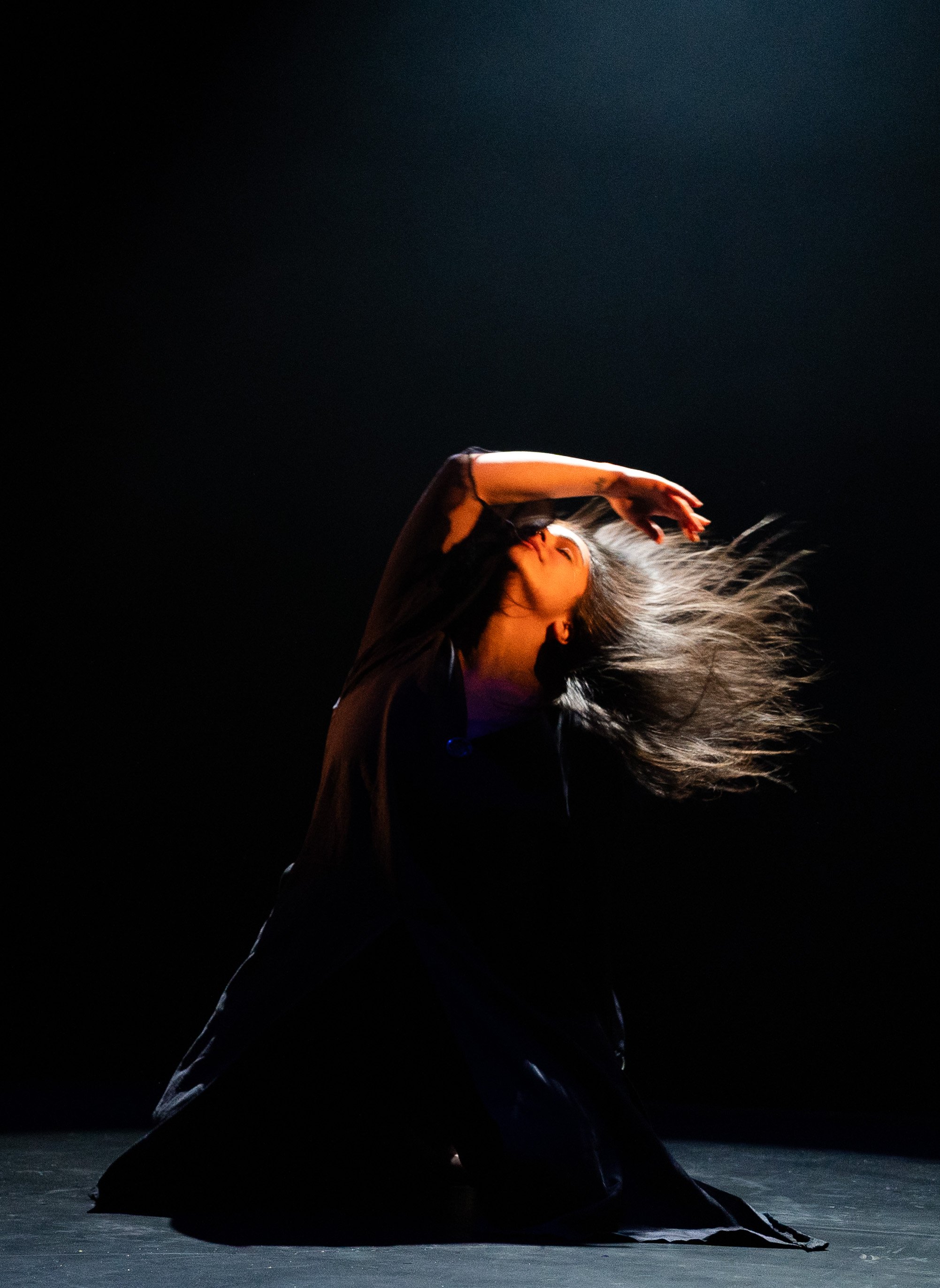 A dancer on stage with long flowing hair, wearing dark clothing, is captured mid-movement with one arm raised above her head and her face turned upward, against a dark background.