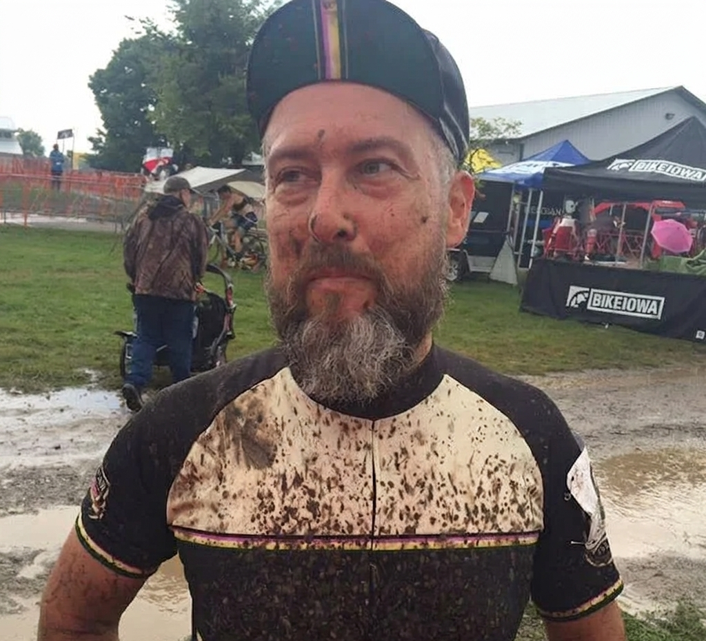 A man in cycling gear, muddy and smiling, wearing a black helmet. In the background, people with umbrellas and tents at an outdoor event.