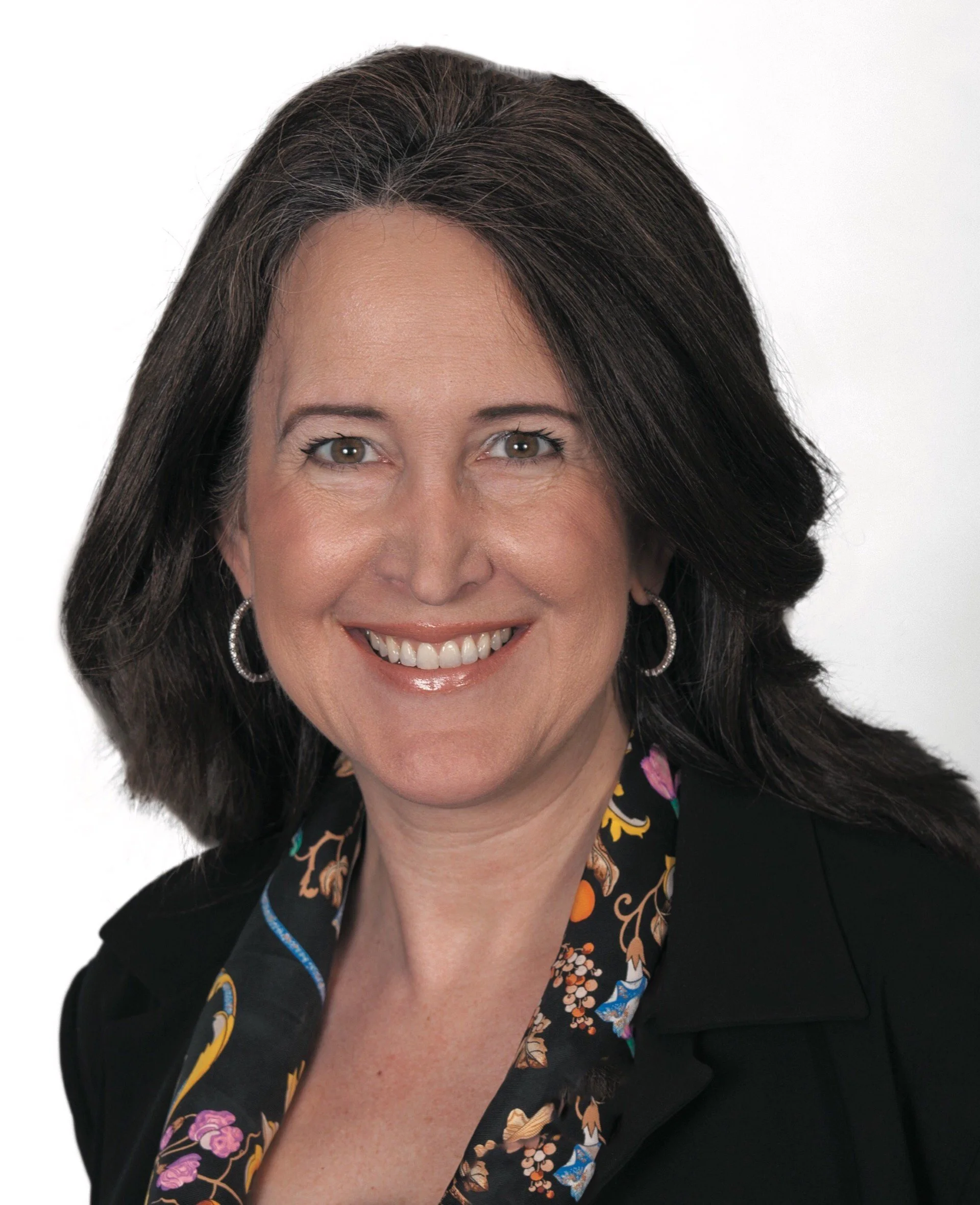 A woman with shoulder-length dark brown hair, wearing hoop earrings, a black blazer, and a floral blouse, smiling against a white background.
