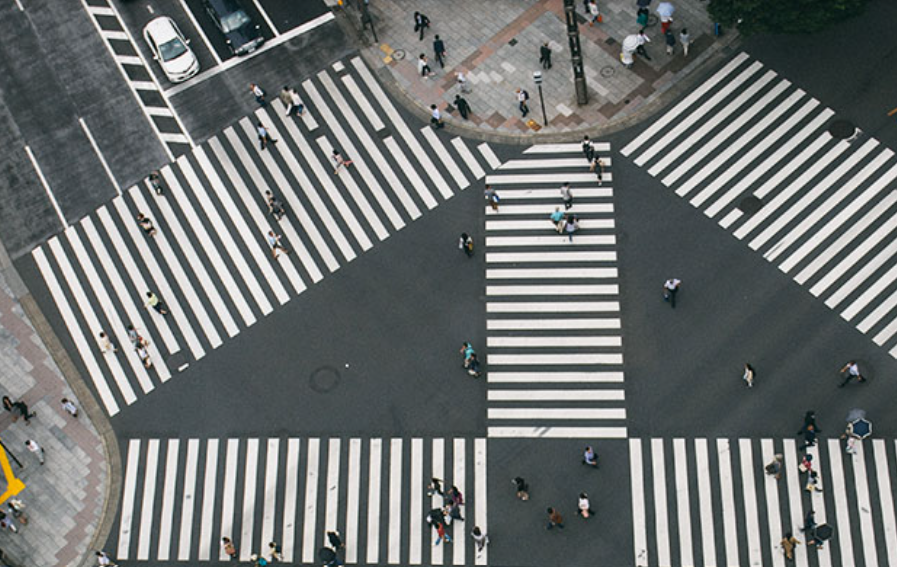 Bird's eye view of a busy city intersection with multiple crosswalks filled with pedestrians and a parking lot with cars.