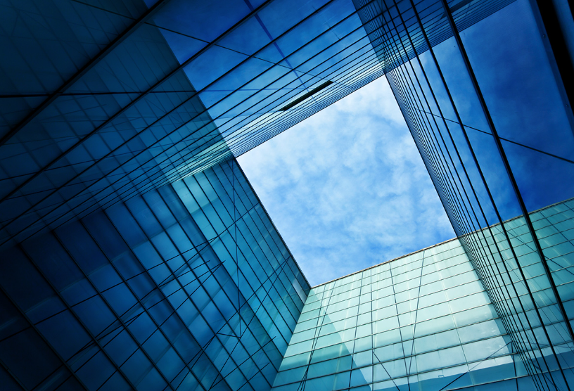 Looking up at the glass facade of a modern skyscraper with a clear blue sky and clouds visible through an open square in the center.