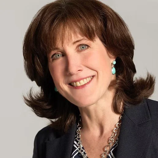 Portrait of a woman with shoulder-length brown hair, blue eyes, wearing turquoise earrings, a dark blazer, and a silver chain necklace, smiling at the camera.