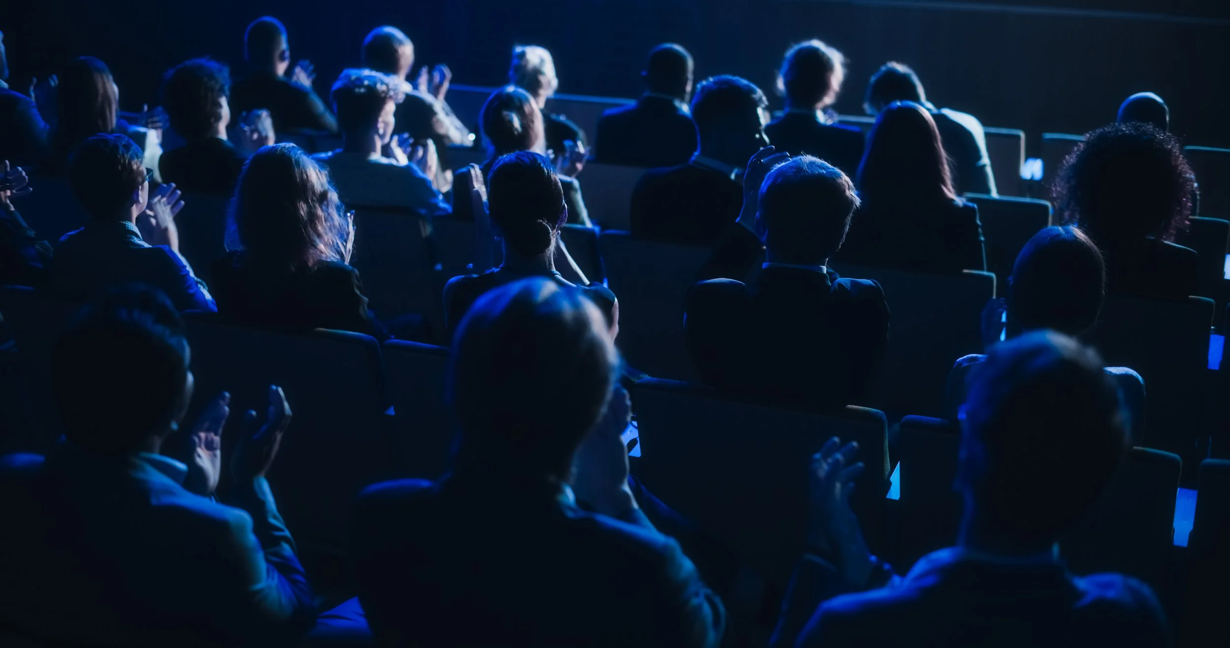 An audience seated in a dark room, watching a presentation or performance on stage with blue lighting.