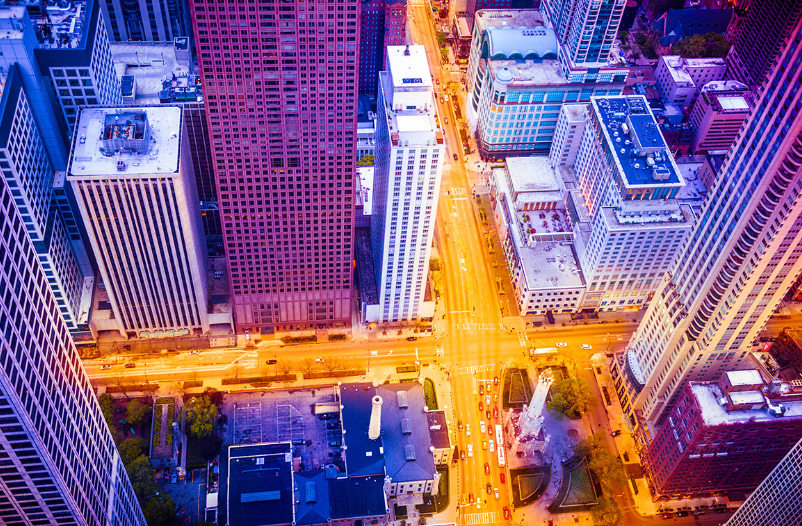 Aerial view of a city block with tall skyscrapers, streets with moving cars, and a small park with a monument.