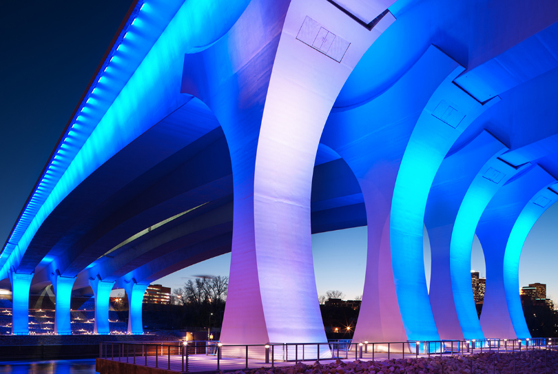 A bridge underpass illuminated with blue and purple lights at dusk, with city buildings visible in the background.