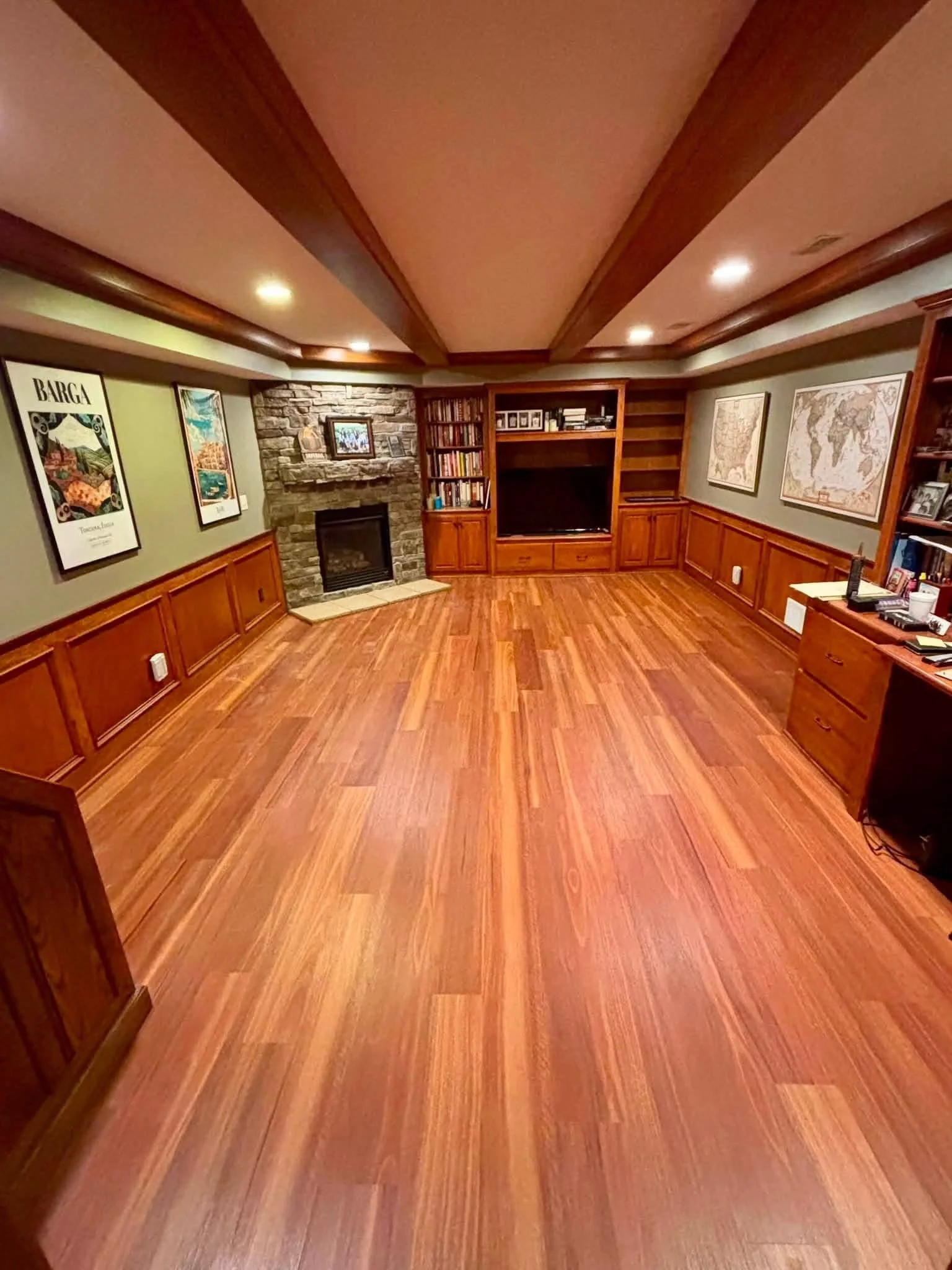 A basement office with cherrywood plank flooring, built in desk, shelves, wainscoting and custom trim, lighting, and a stone fireplace
