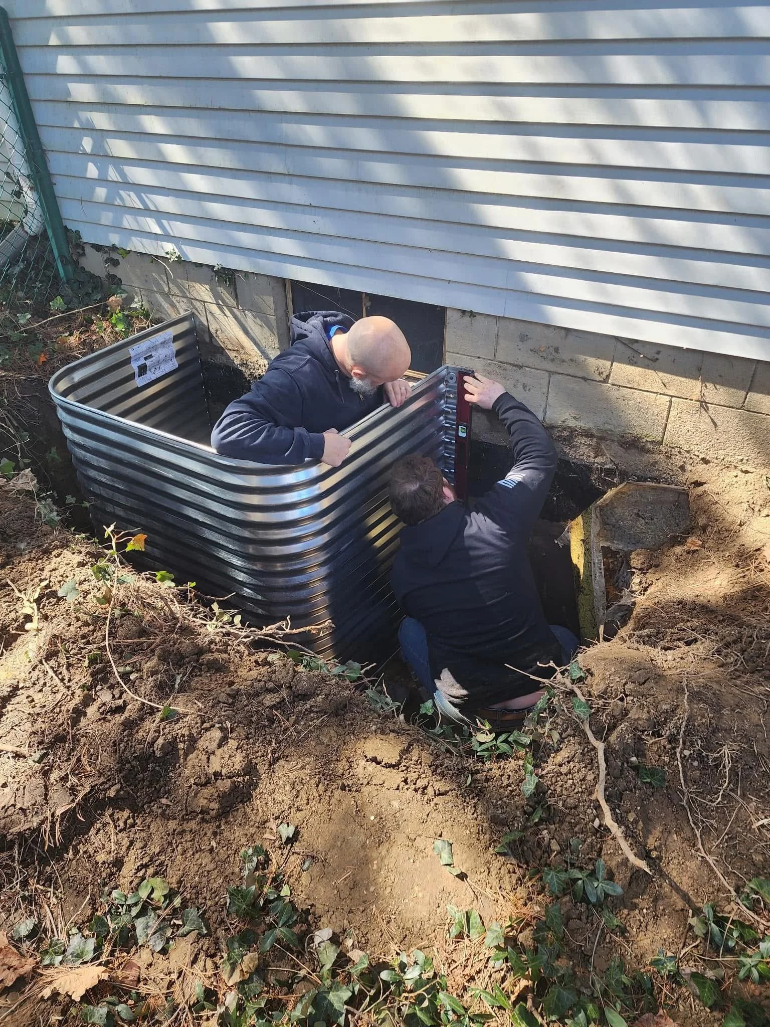Specialists install a metal egress window frame around a basement window