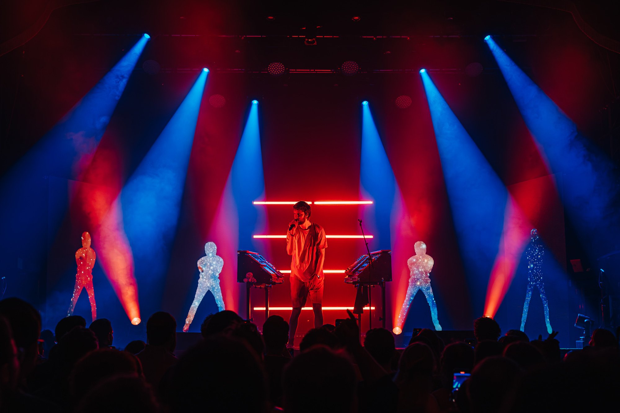 A male performer on stage with vibrant blue and red spotlights, standing in front of digital art mannequins and electronic music equipment, during a live concert.