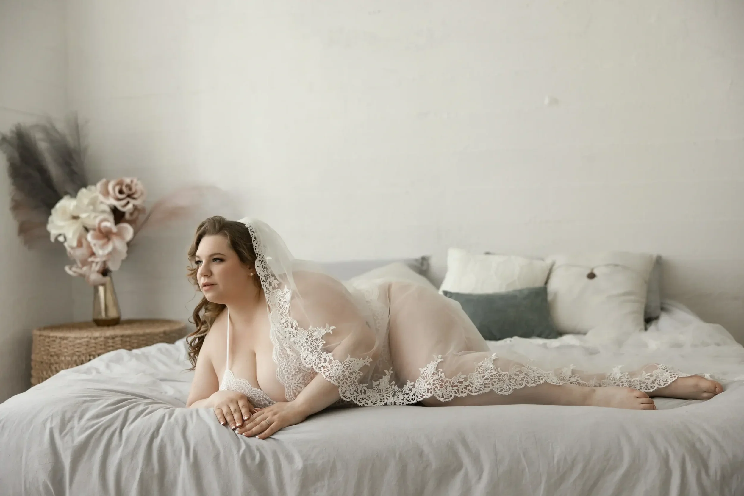 A woman in a sheer, lace-trimmed robe lying on a bed in a well-lit bedroom, with decorative pillows and a floral arrangement in the background posing for her bridal boudoir photo shoot