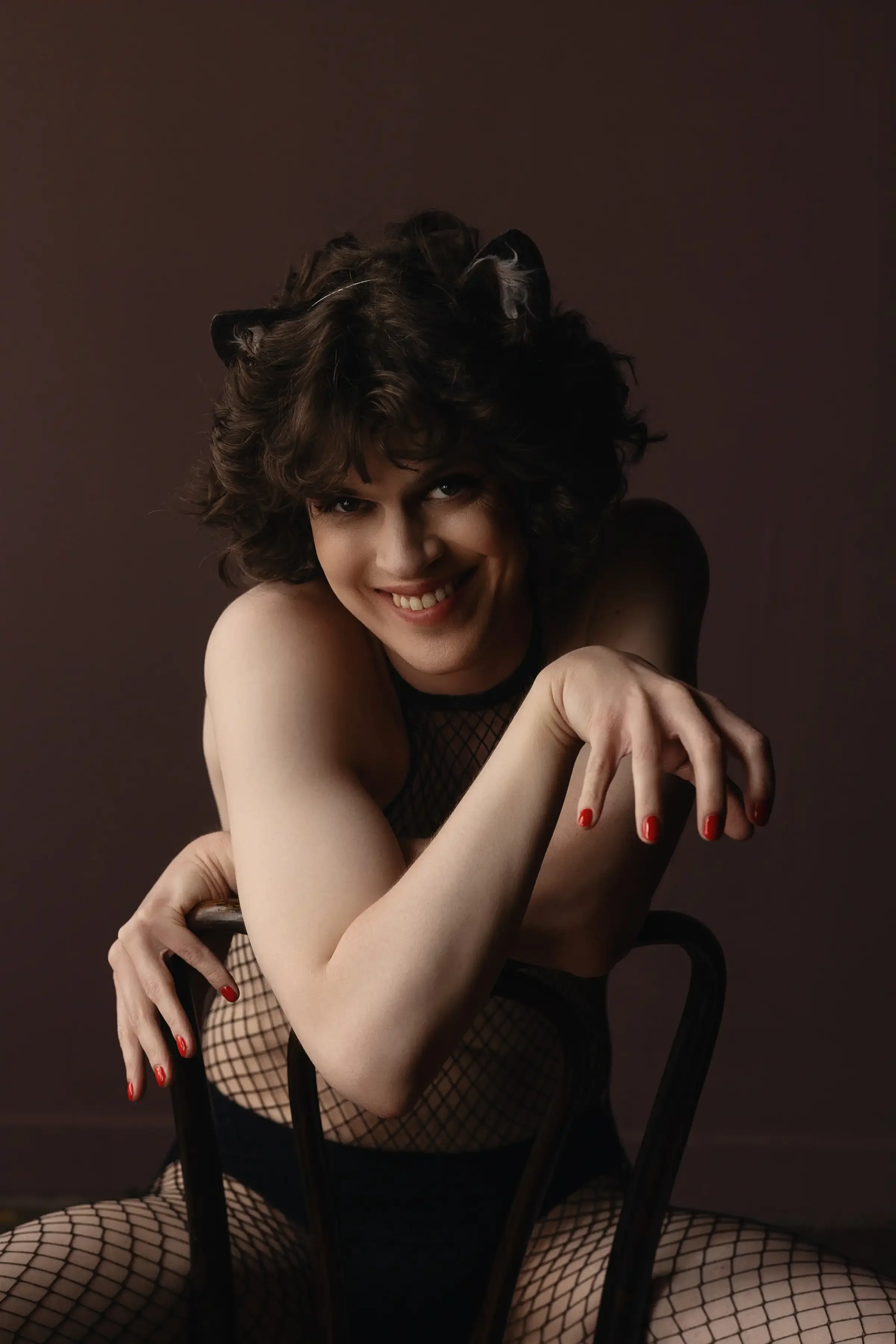 A woman with curly brown hair smiling, wearing fishnet clothing, sitting on the floor with a dark background, leaning on a black chair.