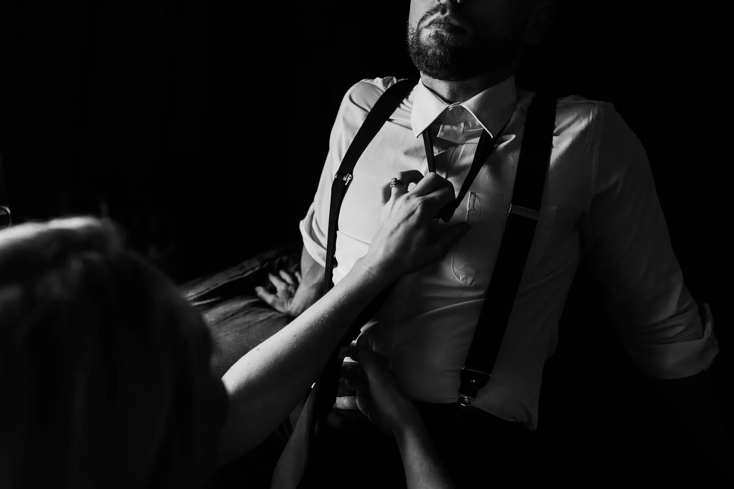 A woman is reaching out and touching the chest of a man wearing a collared shirt and suspenders, sitting with one hand behind him, in a dark, moody scene.