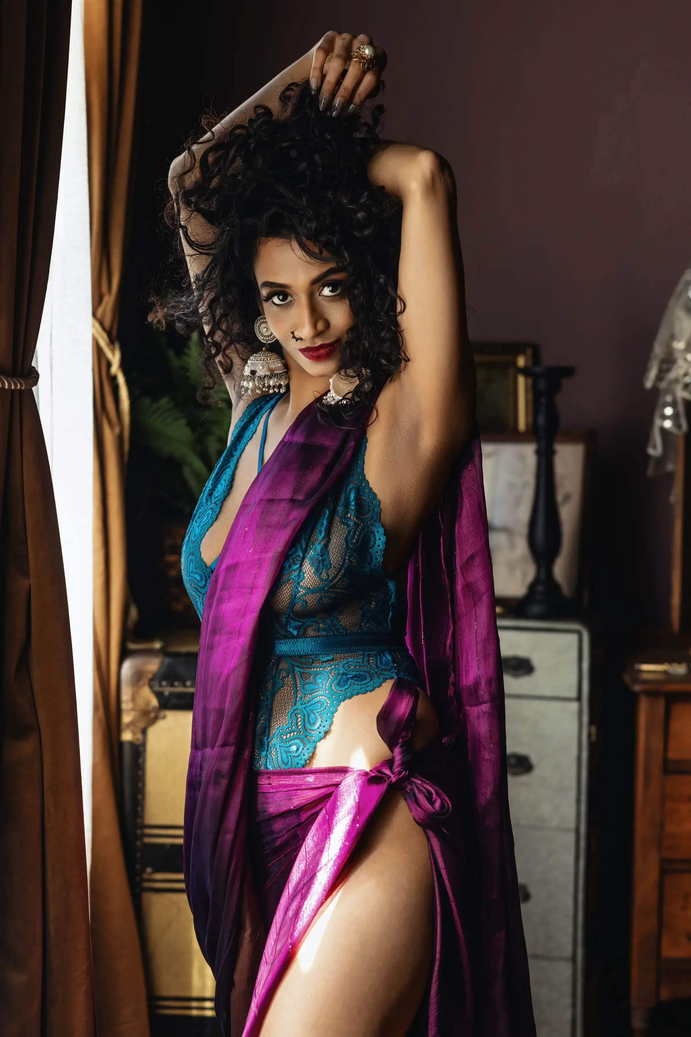 Indian Woman with curly black hair wearing a revealing blue lace lingerie with a purple sheer wrap, standing by a window with brown curtains, posing with her arms raised and smiling at the camera in a dimly lit room.
